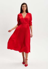 Red pleated midi dress in satin with a V-neckline, long sleeves, and a fitted waist. Paired with matching red strappy high heels.