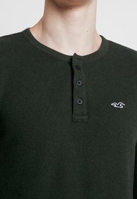 Camisa henley de manga larga en verde oscuro, confeccionada en tela texturizada, con cuello redondo, tres botones negros y un pequeño logo blanco en el pecho.
