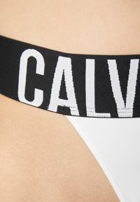 Close-up van een zwarte tailleband met grote witte letters "CALV" op witte ondergoed dat op de huid wordt gedragen.