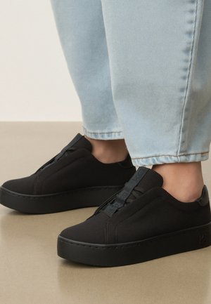 Osoba w jasnoniebieskich dżinsach i czarnych wsuwanych trampkach z grubą podeszwą stojąca na beżowej podłodze.