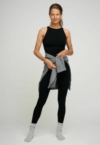 Černý top bez rukávů, černé legíny, šedý pletený svetr svázaný v pase a šedé ponožky. Modelka pózuje na neutrálním pozadí.