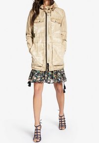 Beżowa parka z kapturem, sznurkiem i kieszeniami z klapką. Noszona na kwiecistej spódnicy, z czarnym i kolorowym wzorem kwiatowym. Sandały na szpilkach.