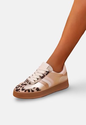 Pied portant une basket beige et marron avec un détail imprimé léopard et des lacets blancs sur une semelle plate en gomme.