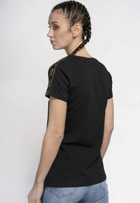 Black cotton t-shirt with a crew neck and short sleeves, featuring gold accent stripes along the shoulder seams. Smooth texture.