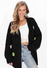 Svart stickad cardigan med överdimensionerade ärmar, blommig brodyr i lila och grönt på ärmarna, och en öppen frontdesign.