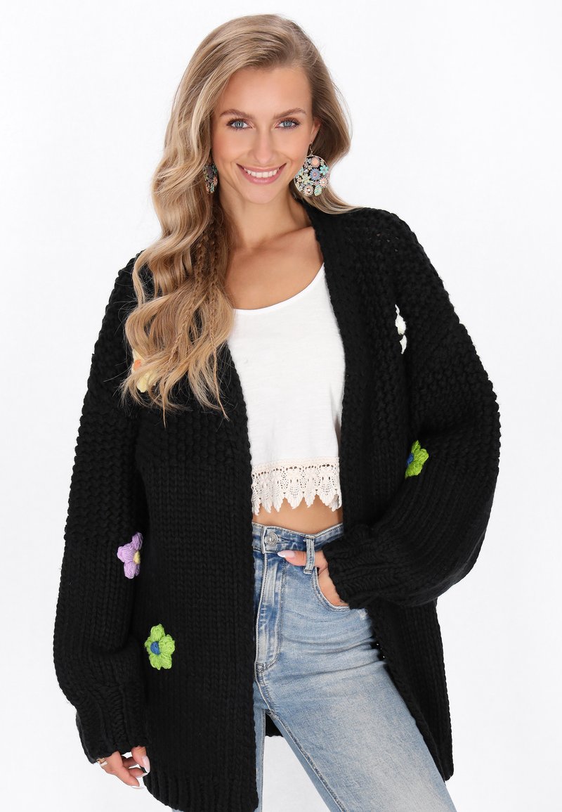 Svart stickad cardigan med överdimensionerade ärmar, blommig brodyr i lila och grönt på ärmarna, och en öppen frontdesign.