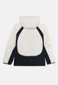 Veste à capuche à manches longues, partie supérieure beige et partie inférieure noire, avec un grand panneau noir incurvé au dos.