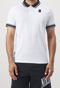 Mies, joka pukeutuu valkoiseen lyhythihaiseen polo-paitaan, jossa on musta kaulus ja hihansuut, yhdistettynä mustiin shortseihin, joissa on iso Nike-logo.