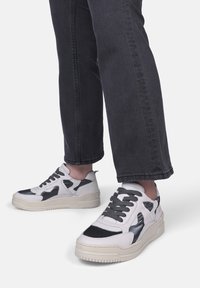 Hvide og sorte sneakers med en tekstureret overflade og en kraftig sål, der har snørebånd og udskæringsdesignelementer. Bæret med mørke jeans.