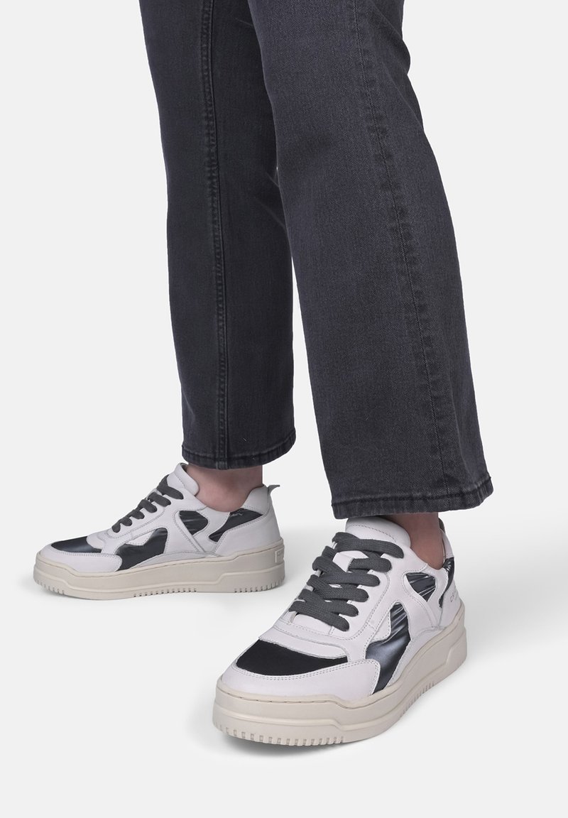 Hvide og sorte sneakers med en tekstureret overflade og en kraftig sål, der har snørebånd og udskæringsdesignelementer. Bæret med mørke jeans.
