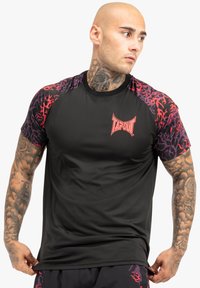 Kortærmet sort sports T-shirt med lilla og røde mønstrede ærmer, forsynet med en rød "TAPOUT" logo på brystet. Glat, letvægtsstof.