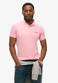 Jasnoróżowa koszulka polo z kołnierzykiem, dwoma guzikami i haftowanym logo. Noszona z niebieskimi dżinsami i szarym swetrem związanym w talii.