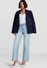 Donna che indossa un blazer oversize blu navy sopra una maglietta bianca, jeans dritti azzurri chiaro e tacchi slingback bianchi appuntiti, in piedi davanti a uno sfondo semplice.