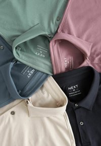 Cinco camisetas tipo polo con cuello en varios colores: verde, marino, rosa pálido, negro y beige. Tejido suave con botones y etiquetas de marca visibles.