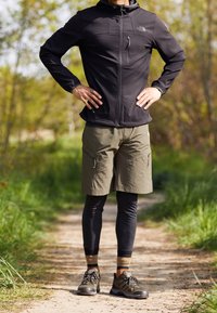 Czarna kurtka na zamek, oliwkowe szorty, czarne legginsy i wytrzymałe obuwie turystyczne na męskiej sylwetce w naturalnym otoczeniu.