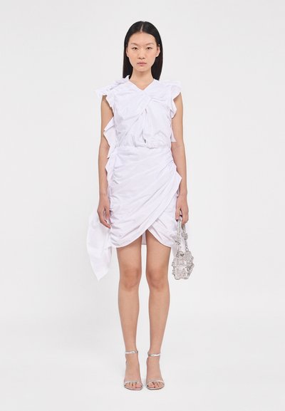Robe blanche avec des manches à volants et un design en tissu torsadé, présentant un ourlet drapé et asymétrique. Associée à un sac à main argenté et à des talons à lanières.