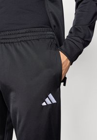 Black athletic pants made from smooth, lightweight fabric. Features an elastic waistband, side pockets, and a grey three-stripe logo detail.