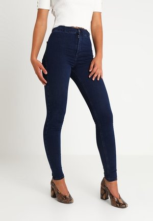 Kobieta w ciemnoniebieskich dopasowanych dżinsach, białym krótki topie i wysokich szpilkach w wzór skóry węża, stojąca na jednolitym tle.