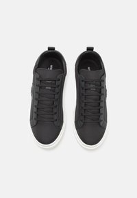 Black sneakers with a textured mesh upper, flat laces, and white rubber sole. Features a logo accent near the heel. Minimalist design.