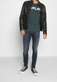 Hombre con chaqueta de cuero negra sobre una camiseta Replay verde oscura, jeans ajustados azules y zapatillas negras con cordones y suelas blancas.