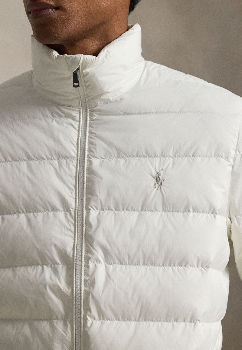 Veste matelassée blanche avec col montant, fermeture éclair et texture matelassée horizontale. Comprend un petit logo brodé sur la poitrine.