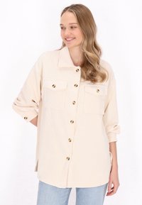 Cămașă bej cu tricot cu butoane de ton auriu și două buzunare pe piept. Cămașa dispune de o guler și mâneci răsucite.