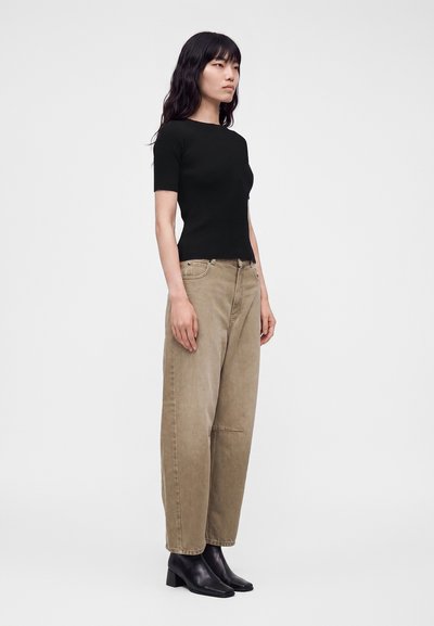 Femme aux longs cheveux noirs portant un haut noir ajusté à manches courtes, un pantalon beige ample et des bottines noires à talons, debout de côté.