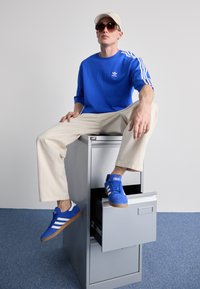 Kék pamut pulóver fehér Adidas logóval, világos bézs széles szárú nadrág, kék velúr sportcipő fehér csíkokkal, és bézs baseball sapka.