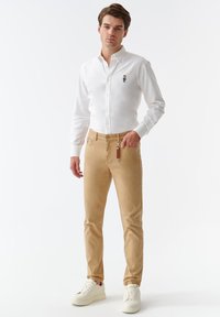 Camisa blanca liviana de botones con detalles bordados, combinada con pantalones ajustados beige. Las zapatillas tienen una suela elevada, lo que proporciona un aspecto casual.