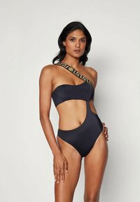 Čierny jednodielny plavky s asymetrickým dizajnom, výrezmi na bokoch a kontrastným vzorovaným popruhom so zlatými akcentmi. Hladká textúra.