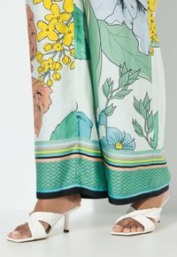 Robe longue à motif floral dans des teintes de vert, jaune et marron ; rehaussée de rayures horizontales à l'ourlet. Une paire de talons blancs à lanières.