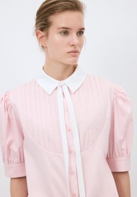 Blusa rosa con un cuello blanco, botonadura frontal y mangas cortas abullonadas. Pliegues verticales adornan la parte superior, confeccionada en una tela suave.