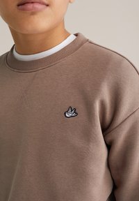 Közeli kép egy törzsről, amely barna pulóvert visel, kis fehér madár emblémával a mellkasán, fehér kerek nyakú póló felett.