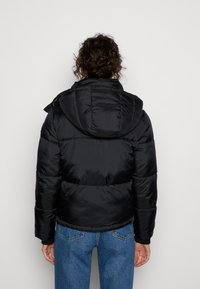 Chaqueta negra acolchada con capucha, con diseño acolchado, puños elásticos y textura suave y brillante; combinada con vaqueros azules.