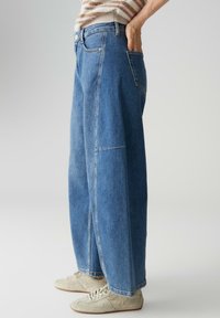Jean en denim bleu clair à jambes larges et taille haute, avec poches appliquées et détails d'ourlet brut, porté avec des baskets de couleur claire.