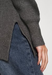 Nahaufnahme einer Hand, die an der Hüfte ruht, trägt einen strukturierten grauen Pullover mit seitlichem Schlitz und blaue Jeans.
