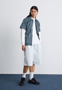 Short-sleeve, teal-striped shirt over a white long-sleeve, paired with light blue shorts and black sneakers. Textured fabric, relaxed fit.