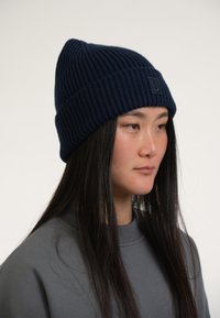 Gorro de punto azul marino con textura acanalada y un pequeño parche con logo, usado por una persona con cabello largo, combinado con una sudadera gris.