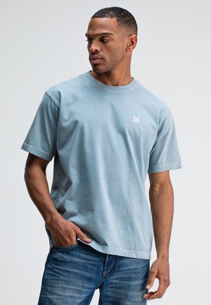 Lichtblauwe katoenen T-shirt met een ronde hals, korte mouwen en een vierkant logo op de borst. Draagt met blauwe jeans, met een relaxed fit.