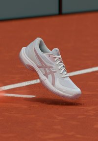 Scarpa sportiva bianca e rosa che solleva argilla rossa su un campo da tennis vicino a una linea di confine bianca.