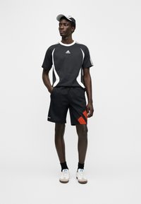 Svart sport-t-shirt med vita detaljer, kombinerad med svarta shorts med röda diagonala ränder. Modellen bär vita skor och keps.