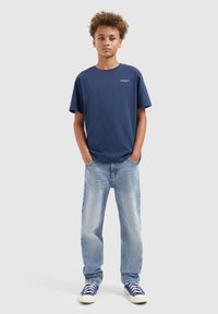 Tienerjongen die staat met zijn handen in zijn zakken, in een marineblauw t-shirt, lichtblauwe jeans en blauwe sneakers, tegen een effen grijze achtergrond.