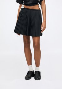 Μαύρη πλισέ φούστα, κατασκευασμένη από ελαφρύ ύφασμα, διαθέτει ελαστική μέση. Φοριέται με μαύρα αθλητικά παπούτσια και λευκές κάλτσες πλήρους μήκους.