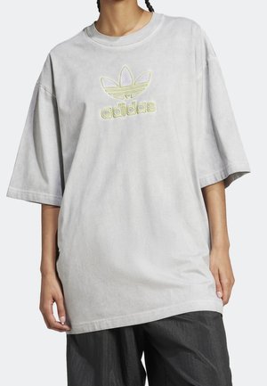 Personne portant un t-shirt Adidas gris clair surdimensionné avec un logo brodé jaune néon et un pantalon noir ample, debout les mains détendues.