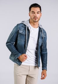 Jeansjacke in verblasstem Blau mit grauem Kapuze, ausgestattet mit vorderen Taschen und einem Reißverschluss, getragen über einem weißen T-Shirt und beigefarbenen Hosen.