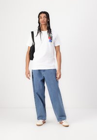 White cotton t-shirt with printed colorful design on left chest, paired with light blue wide-leg jeans and cream sneakers with red stripes.