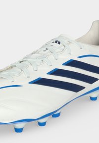 Λευκό ποδοσφαιρικό παπούτσι με μπλε ρίγες Adidas, μπλε τρακτέρ στη σόλα και λευκά κορδόνια σε απλό φωτεινό φόντο.