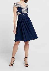 Mörkblå klänning med korta ärmar, blommig spetsdetalj, en figurnära överdel och en utsvängd kjol, i kombination med svarta högklackade sandaler.