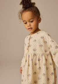 Beige Kleid mit floralem Muster, langen Ärmeln, Rundhalsausschnitt und Holzknöpfen. Verfügt über kleine Punkte und Fältchen an der Taille.