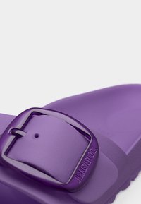 Κοντινό πλάνο στο λουράκι ενός μοβ σανδαλιού Birkenstock με λαμπερή μοβ αγκράφα και ανάγλυφο όνομα της μάρκας.